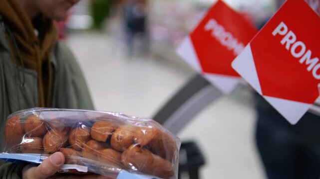 Shopper holds packaged bread beside bright promo signs in grocery store. Budget minded customer compares marked down food, considering affordable purchase while browsing supermarket shelves.