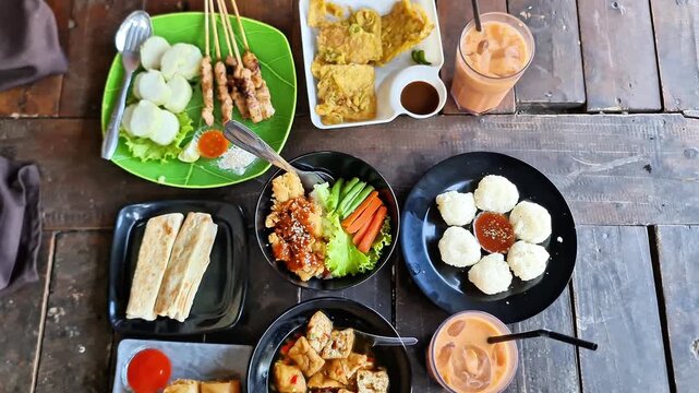 Top View of Authentic Indonesian Culinary Feast Featuring Chicken Satay, Cireng Rujak, Fried Tofu, and Iced Tea on a Rustic Wooden Table