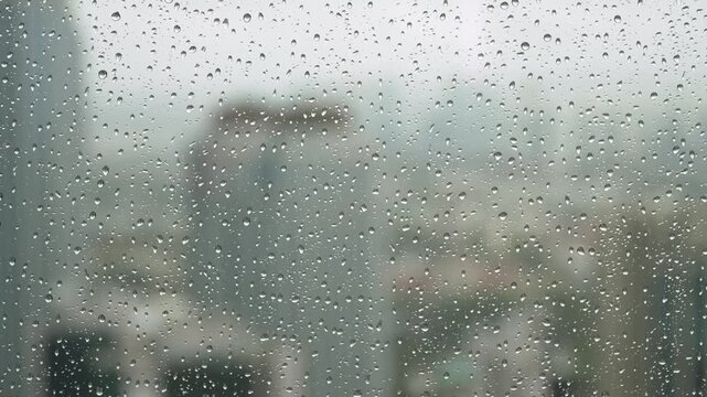 Raindrops on window glass with a blurred city skyline of high-rise buildings behind, a gray rainy day seen from indoors, weather background