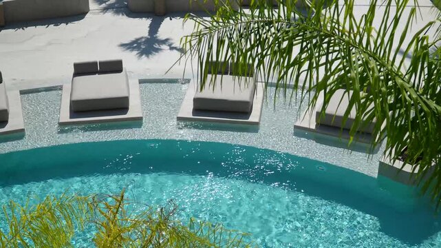 Luxury resort swimming pool with turquoise water and sunbeds on an in-pool ledge, framed by palm fronds, high angle view of a hotel pool on a sunny summer day