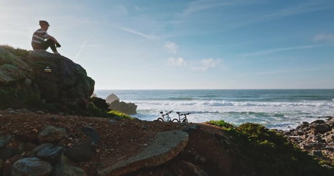 Man in cycling gear resting on rock enjoying the scenic Atlantic Ocean view with two mountain bikes parked nearby on the rugged coastline, adventure, freedom, and an active lifestyle. Portugal, Lisbon