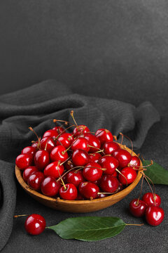 Fresh ripe red sweet cherries in wooden bowl on dark textured stone background with green leaves and black linen towel. Summer fruit, berry, harvest, healthy organic food