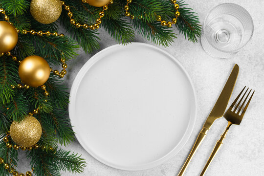 Christmas table setting with empty white plate, golden cutlery, green spruce branch with gold balls, on gray background. Template, mockup with festive aesthetic holiday atmosphere.