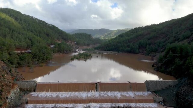 Namnam River reservoir in Koycegiz, Mugla, Turkey, aerial drone view flying from the stepped weir over muddy floodwater reflecting clouds between pine-forested hills