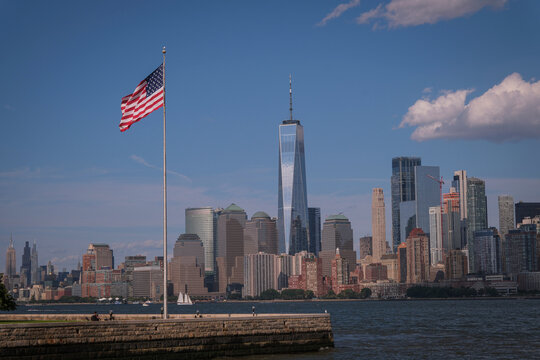 American flag on the jetty of Liberty Island as a sailing boat passes in front of Lower Manhattan, New York City, United States