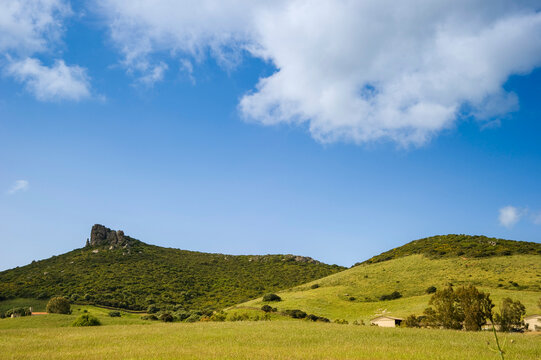 landscape with mountains and blue sky, a rolling green rural landscape with a rocky outcrop on a hill under a blue sky with white cumulus clouds, consistent with the Sardinian Countryside in Italy