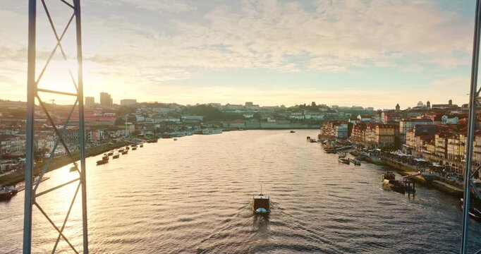 Douro River at sunset winding through Porto historic waterfront with traditional rabelo boats beneath the iconic Luis I Bridge, warm golden reflection on the water. Aerial drone fly inside arch window
