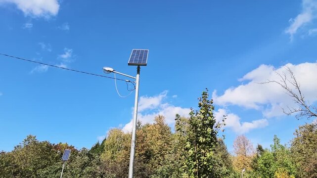 Solar panel street lamp on a metal pole against a blue sky above autumn trees.