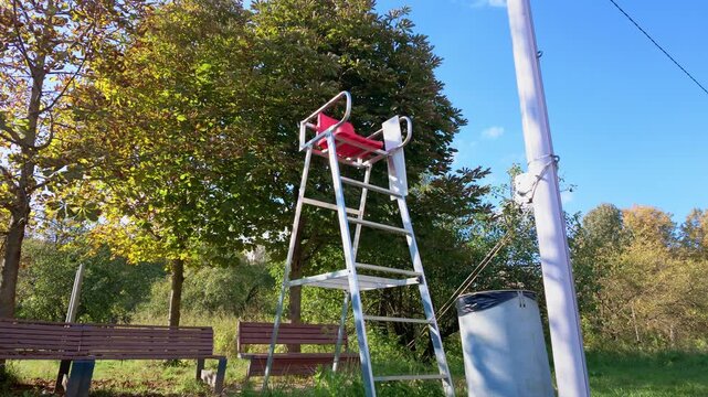 Empty aluminum referee chair with red seat beside benches in a sunny autumn park.
