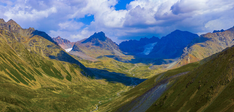 Panoramic view captures a mountain valley featuring a glacier in the background. Rolling green hills and a stream flow through the valley floor under a partly cloudy sky.