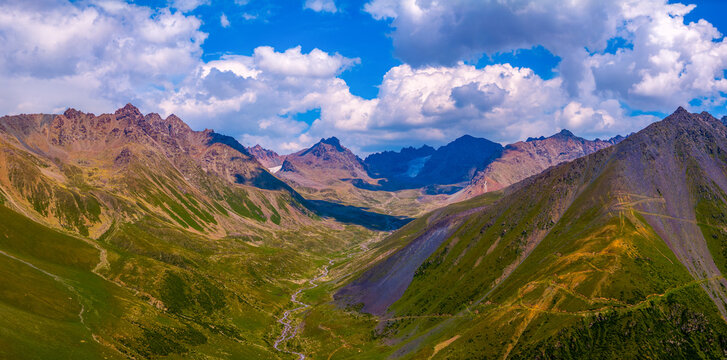 Aerial perspective captures a mountain valley featuring a meandering stream. Steep slopes covered in green vegetation rise on either side, with distant jagged peaks visible under a partly cloudy sky.