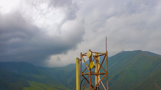 Telecommunications tower with circular antennas stands prominently in front of rolling green mountains. Weathered metal structure contrasts with lush vegetation under a cloudy sky.
