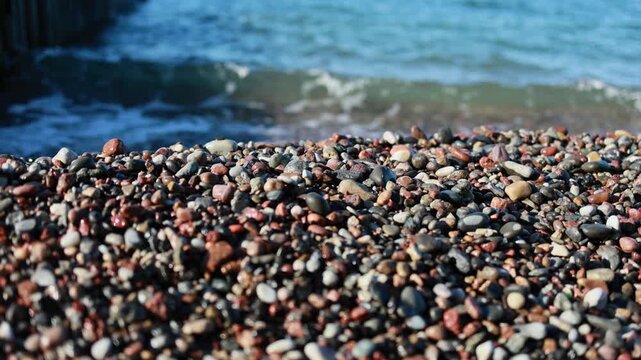 Waves rolling onto colorful pebble beach on seashore. Smooth sea stones close up with blue water surf in background. Nature coastal landscape of rocky beach