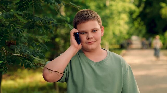 boy enjoying call outdoors, teen boy talking on smartphone, talking cheerfully along forest path today, cheerful boy on voice call, standing on pathway in green park, green trees in background, green