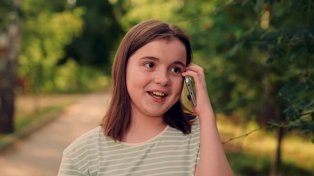 girl on phone, relaxed smile, greenery blur in background., smiling youngster enjoys friendly conversation on smartphone in parkway today., young girl with striped shirt talks on phone, teen girl