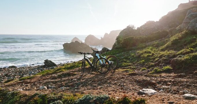 Two bicycles parked on rugged, sunlit cliffside path, offering a panoramic view of Atlantic ocean's waves crashing against a rocky coastline near Lisbon, Portugal. Outdoor activity, sport, recreation