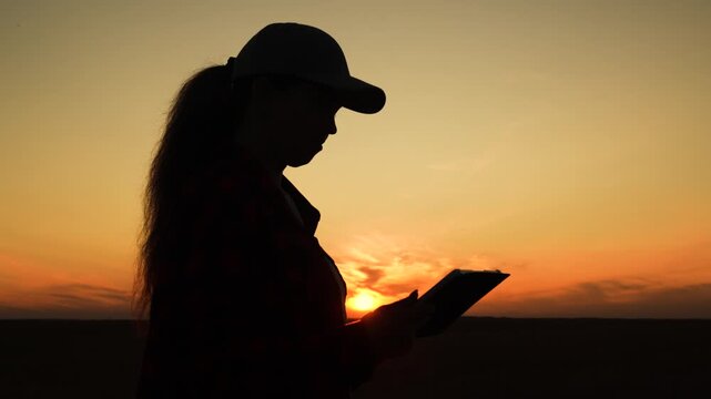 Silhouette woman working digital tablet, young girl sunset, human journey digital technology, cool tech in the countryside, reading the screen as the sun goes down, golden hour light outlines a person