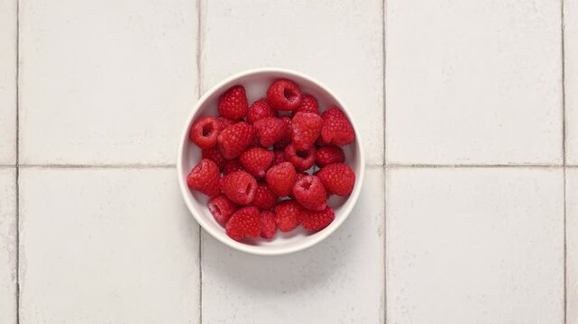 Hands placing a bowl of fresh raspberries on a white tiled kitchen table and picking one
