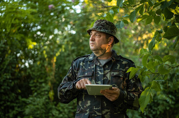 Man in camouflage uniform consults tablet while standing among forest greenery. Alert soldier holds screen with both hands, maintaining focused expression beneath overhanging leaves. © Віталій Б.