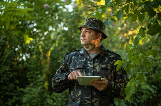 Man in camouflage uniform consults tablet while standing among forest greenery. Alert soldier holds screen with both hands, maintaining focused expression beneath overhanging leaves.