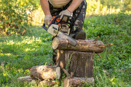 Man uses chainsaw to cut trunk supported by stump in grassy woodland. Wearing gloves and camouflage trousers, focused worker prepares timber with power equipment outdoors, guiding saw through trunk.