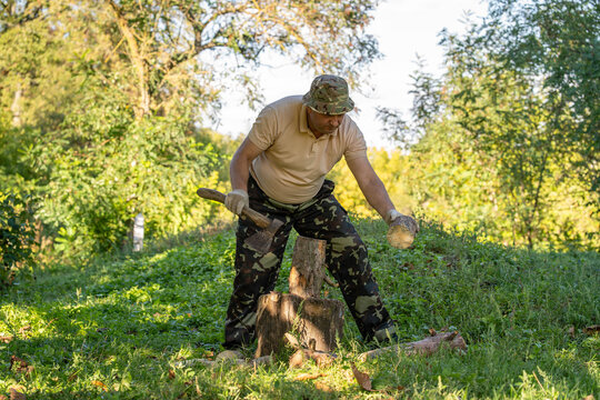 Man splits firewood outdoors, holding log beside chopping stump and gripping axe. Focused woodcutter wears camouflage trousers and bucket hat in sunlit green woodland clearing, working with hand tool.