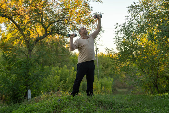 Man exercises with dumbbells outdoors, holding one weight overhead and another near shoulder. Focused fitness enthusiast stands among green trees and grass during strength training in nature.