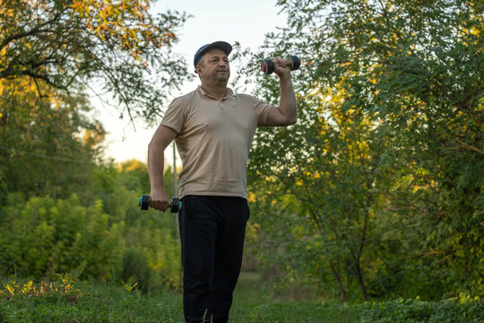 Middle aged man holds dumbbells outdoors, raising one weight beside his shoulder while other hangs at his side. He looks focused during strength training on leafy woodland path, working his arms