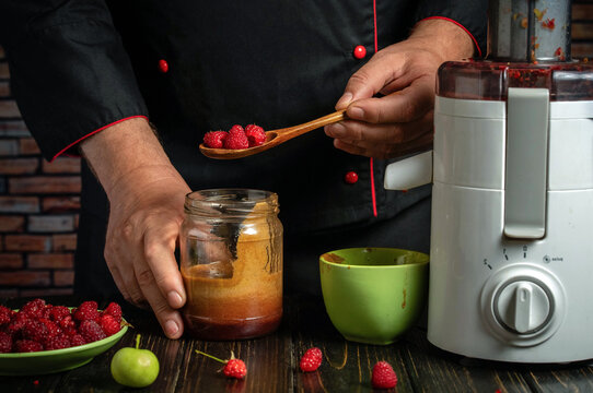 Chef holds raspberries on wooden spoon beside blender and jar of puree. Black uniform with red trim frames fresh berries prepared for homemade fruit smoothie in rustic kitchen, with berries and puree.