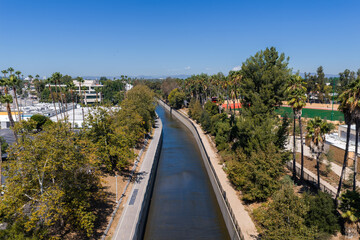 Fototapeta premium Ballona Creek concrete flood control channel flows beside a paved bicycle path and trees under a clear sky in Culver City, Los Angeles County, California.