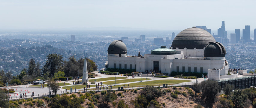 Griffith Observatory sits on the slope of Mount Hollywood in Griffith Park, with visitors on the grounds and the downtown Los Angeles city skyline visible under hazy daylight.