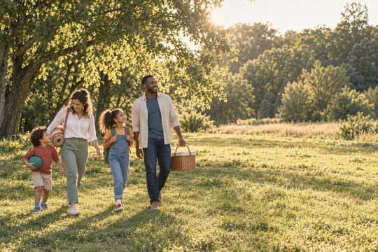 Family of four walking through a sunlit park with picnic supplies