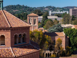 Fototapeta premium Romanesque revival brick architecture of UCLA campus shows tile roofs and bell towers framed by forested hills of Los Angeles under clear daytime skies.
