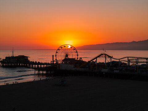 Santa Monica Pier Ferris wheel and roller coaster are silhouetted against a golden sunset over the Pacific Ocean, with distant mountains along the California coastline.