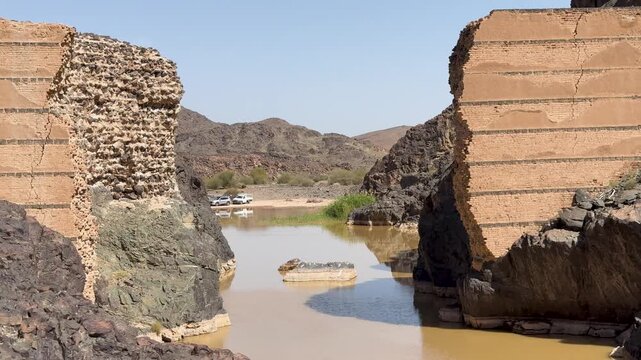 Broken ancient dam ruins with muddy water in a rocky canyon in Saudi Arabia.