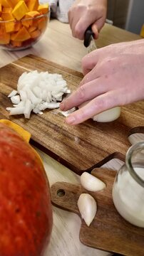 A person uses a knife to chop a white onion on a wooden cutting board, with bowls of orange vegetables and garlic cloves nearby on a table