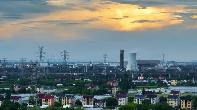 Industrial power plant and cooling tower dominate suburban landscape under dramatic sunset clouds in high angle view.