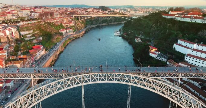 Dom Luis I Bridge connecting Vila Nova de Gaia and Porto, featuring pedestrians on the upper deck, vehicles on the lower deck, and city architecture along the Douro River at sunset. Aerial drone shot