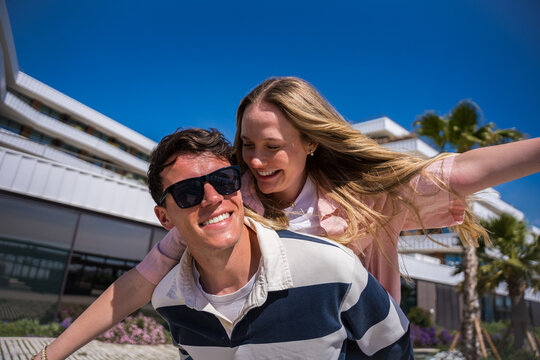 Happy young couple enjoy a playful piggyback ride outdoors in a modern city, laughing together while the woman stretches her arms out under a vivid blue sky.