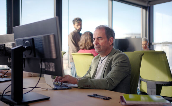 Focused mature businessman typing on a laptop at his desk while colleagues work in the background. Bright corporate scene conveying concentration, responsibility and productivity.