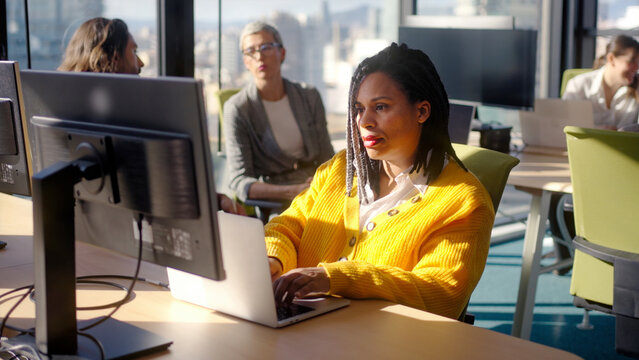 Focused Black businesswoman typing on a laptop at her desk while colleagues work in the background. Sunlit corporate scene conveying productivity, concentration and modern office life.