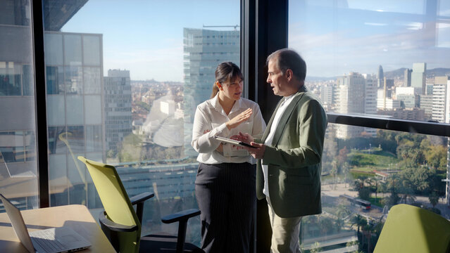 Two businesspeople are standing in a modern office with large windows, overlooking a cityscape, engaging in a discussion while reviewing information on a tablet