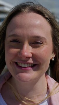 Vertical close up portrait of a happy young White blonde woman smiling directly at the camera as her hair moves in the breeze.