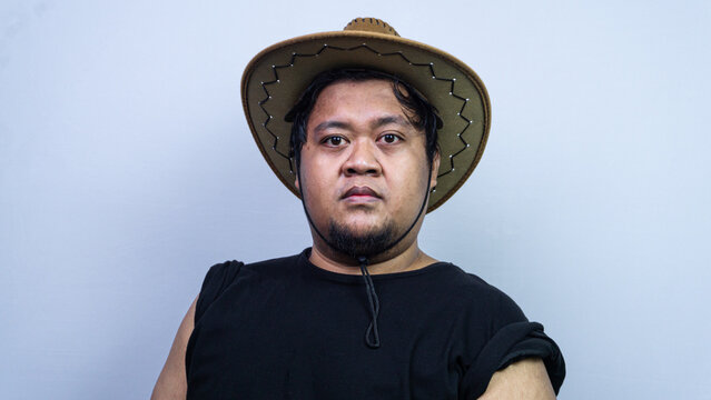 A young Asian male wearing a rustic brown cowboy hat and a dark casual shirt stares directly into the camera with a completely deadpan expression. This minimalist studio portrait focuses