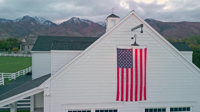 Aerial view of a large American flag hanging on a white barn with autumn mountains in the background