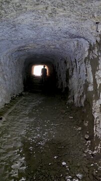 Backlit view of person walking along rock tunnel towards bright illuminated exit arch. Atmospheric footage showcasing hope, exploration, freedom, and finding way out of darkness. Cinematic storytellin
