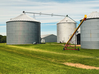 Large metal grain silos, auger equipment, and barns set in a green field under a summer sky on a rural farm near Saugatuck, Michigan. © Sanya Kushak