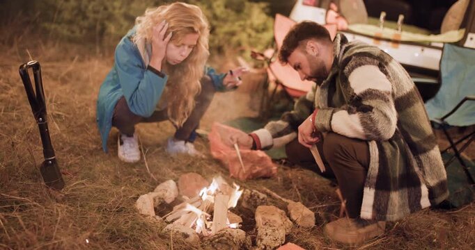Couple cooks food by campfire in the woods while enjoying a night under the stars with friends around the car and tents in the background
