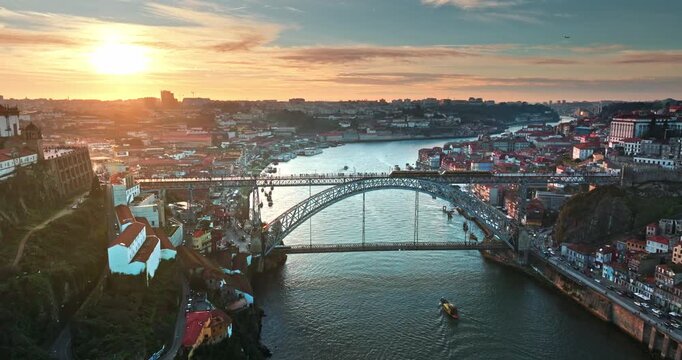 Porto historic city center illuminates at golden hour sunset, iconic Dom Luis I Bridge spanning Douro River, a train crossing the upper deck, traditional boats navigating the water. Aerial drone shot