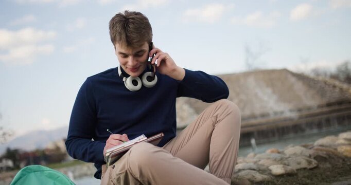 Teenage Boy Multitasking: Studying and Talking on Phone by Lake in Park